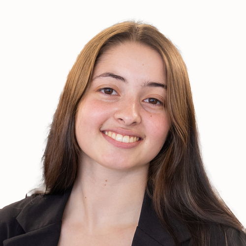 A close-up headshot of a smiling young woman with long brown hair, parted at the center, against a plain light background. She is wearing a dark blazer and looking directly at the camera.